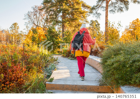Woman walking on wooden path in autumn park. Middle aged female explorer with backpack and camera tripod enjoying fall nature. Outdoor activity, leisure, travel and lifestyle concept. Woman walking on wooden path in autumn park. Middle aged female explorer with backpack and camera tripod enjoying fall nature. Outdoor activity, leisure, travel and lifestyle concept. 140765351