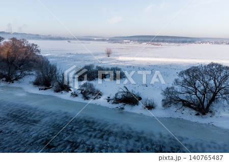 A tranquil winter morning unfolds along a frozen river, with bare trees silhouetted against a pale sky, their branches dusted with frost, creating a serene, frosty landscape A tranquil winter morning unfolds along a frozen river, with bare trees silhouetted against a pale sky, their branches dusted with frost, creating a serene, frosty landscape 140765487