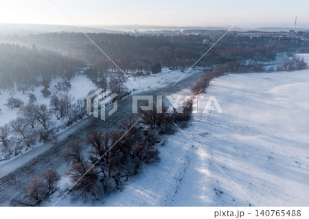 Aerial view of a serene winter river at dawn, framed by snow-covered banks and bare trees, with a hint of distant mist and a hazy horizon Aerial view of a serene winter river at dawn, framed by snow-covered banks and bare trees, with a hint of distant mist and a hazy horizon 140765488
