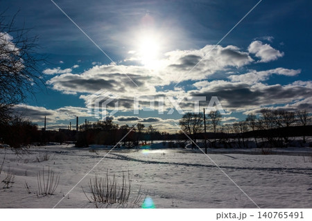 Sunlight pierces through dramatic clouds, illuminating a vast snow-covered landscape with scattered trees and distant industrial structures, a true winter's stark beauty Sunlight pierces through dramatic clouds, illuminating a vast snow-covered landscape with scattered trees and distant industrial structures, a true winter's stark beauty 140765491