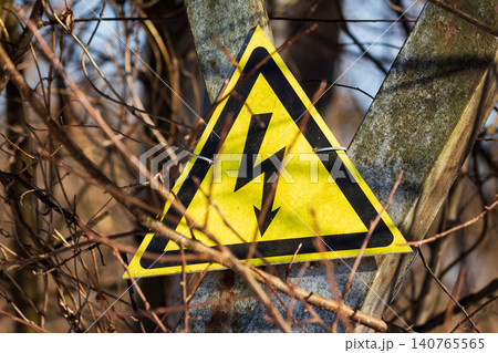 Bright yellow high voltage danger sign mounted on a metal pole surrounded by tangled dry tree branches providing a clear warning symbol outdoors during the cold season 140765565