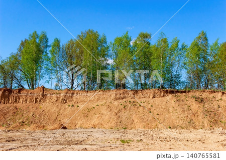 Vivid spring scene of an old sandy quarry cliff face under a brilliant blue sky. Fresh green trees crown the textured earth Vivid spring scene of an old sandy quarry cliff face under a brilliant blue sky. Fresh green trees crown the textured earth 140765581
