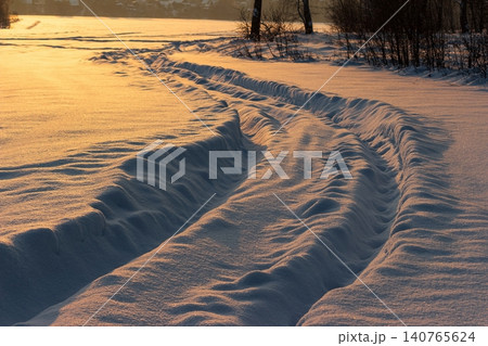 Deep tire tracks carve through pristine snow at sunset, bathed in golden light. A tranquil winter evening landscape 140765624