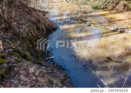 A steep, mossy bank slopes down to a flooded gully, its muddy waters reflecting bare branches and the April sky. A natural scene showcasing the power of spring thaw and rising water levels A steep, mossy bank slopes down to a flooded gully, its muddy waters reflecting bare branches and the April sky. A natural scene showcasing the power of spring thaw and rising water levels 140765629