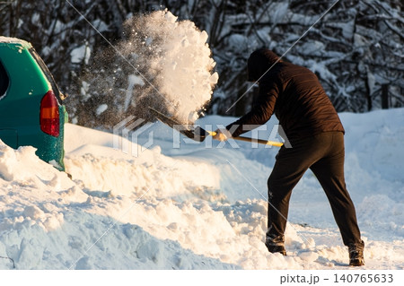 Man actively clearing a substantial snowdrift with a shovel, freeing his car after heavy winter snowfall. A common, demanding winter chore 140765633