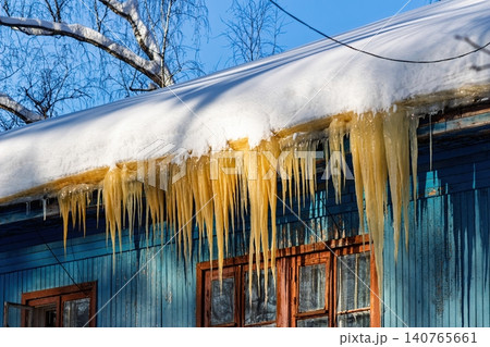 Eerie brownish icicles hang from a snow-laden roof, catching bright sun against an old blue wooden house, creating a striking winter contrast, Russia Eerie brownish icicles hang from a snow-laden roof, catching bright sun against an old blue wooden house, creating a striking winter contrast, Russia 140765661