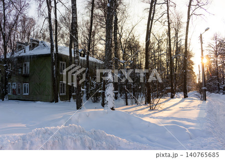 Quaint green wooden house, roof laden with snow and sharp icicles. Bare trees line a snowy path, bathed in the soft glow of a low winter sun, Russia 140765685