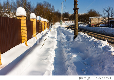 Winter sidewalk scene with deep snow, bright yellow brick pillars, and a metal fence under a crisp blue sky 140765694