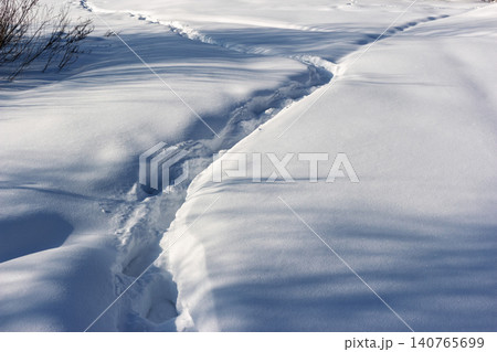 Deep footprints carve a winding path through pristine, sunlit snowdrifts casting sharp blue shadows across the winter landscape 140765699