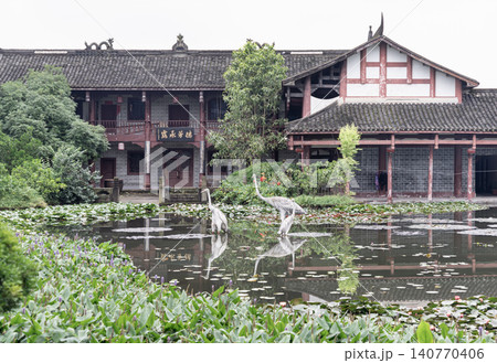 Before the weathered tile roofs and traditional wooden balconies of a grand Chinese estate, a glassy pond rests in perfect stillness. Flanked by lush foliage and floating lotus pads, a group of Before the weathered tile roofs and traditional wooden balconies of a grand Chinese estate, a glassy pond rests in perfect stillness. Flanked by lush foliage and floating lotus pads, a group of 140770406