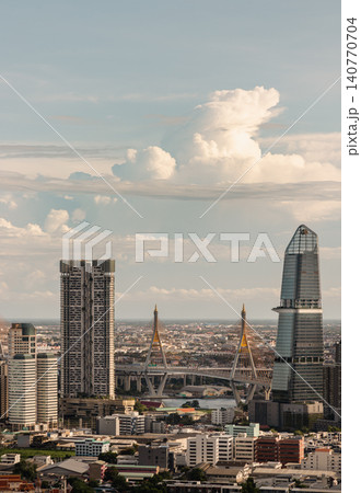 Bathed in warm afternoon light, the city skyline unfolds under a pale sky dominated by a massive, sculpted cumulus cloud. On the right, a sleek, modern skyscraper tapers into a distinctive angled Bathed in warm afternoon light, the city skyline unfolds under a pale sky dominated by a massive, sculpted cumulus cloud. On the right, a sleek, modern skyscraper tapers into a distinctive angled 140770704