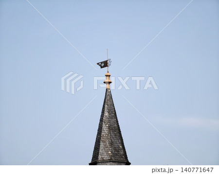Pointed tower with fluttering pennant. Tall conical tower adorned with fluttering vane and distant rooftops Pointed tower with fluttering pennant. Tall conical tower adorned with fluttering vane and distant rooftops 140771647
