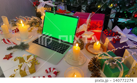 Festive New Year's table with a laptop with a chroma key on the screen. Colorful gift boxes, burning candles, cones, confetti and other elements of Christmas. 140772274
