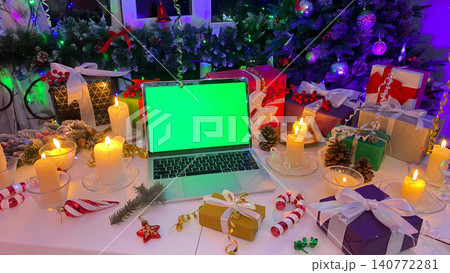 Table decorated with New Year's gifts, burning candles, cones, confetti, fir branches and striped caramel, on the background of a Christmas tree. An open laptop with a chroma key 140772281
