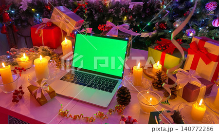New Year's table of a remote worker. Personal laptop with a chroma key on the screen. Bright gift boxes, lighted candles, cones, confetti and other elements of Christmas. 140772286