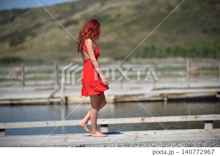 Woman with red hair in red dress enjoying peaceful poolside scenery on a sunny day 140772967