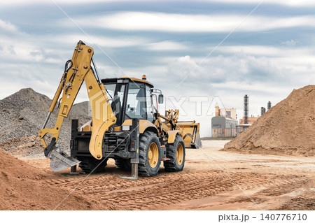 A universal excavator loader digs into the ground at a construction site near buildings and mounds of sand and dirt under a cloudy sky A universal excavator loader digs into the ground at a construction site near buildings and mounds of sand and dirt under a cloudy sky 140776710