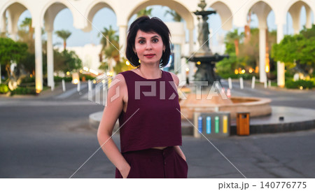 Woman standing in front of an elegant fountain with a grand arched entrance in the background 140776775