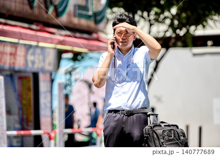 屋外でスマートフォンで通話しながら日差しを遮る夏バテの仕事中の半袖の30代の日本人の男性の会社員 屋外でスマートフォンで通話しながら日差しを遮る夏バテの仕事中の半袖の30代の日本人の男性の会社員 140777689