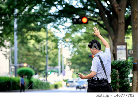 通りでアプリで配車したタクシーを呼ぶためにスマートフォンを持って手を挙げる30代の日本人の男性の会社 通りでアプリで配車したタクシーを呼ぶためにスマートフォンを持って手を挙げる30代の日本人の男性の会社 140777702
