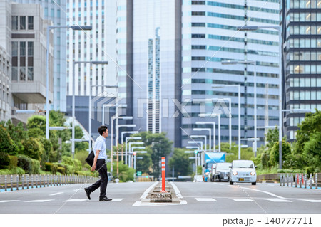 都会の道路の横断歩道を歩く半袖シャツを着た30代の日本人の男性の会社員と高層ビル群の背景 都会の道路の横断歩道を歩く半袖シャツを着た30代の日本人の男性の会社員と高層ビル群の背景 140777711