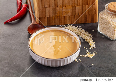Sesame sauce in white bowl with scattered seeds red chili peppers wooden spoon and cutting board on dark surface 140780167