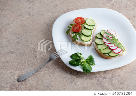 Sandwich with cottage cheese, cucumbers and cherry tomatoes and radish on a white plate on the table. Plate with a fork on the table with a white plate with a salad of vegetables. Sandwich with cottage cheese, cucumbers and cherry tomatoes and radish on a white plate on the table. Plate with a fork on the table with a white plate with a salad of vegetables. 140781294