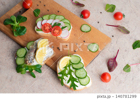 A set of sandwiches with sprats,  homemade curd cream with herbs, with radishes, cucumbers, and tomatoes, on a wooden background. Top view. Copy space. 140781299