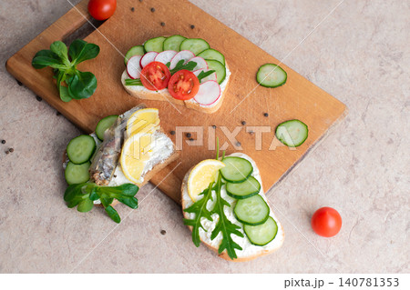 A set of sandwiches with sprats, homemade curd cream with herbs, with radishes, cucumbers, and tomatoes, on a wooden background. Top view. Copy space. A set of sandwiches with sprats, homemade curd cream with herbs, with radishes, cucumbers, and tomatoes, on a wooden background. Top view. Copy space. 140781353