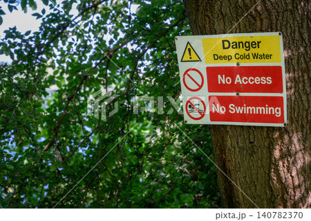 Danger sign warning of deep cold water. No access. No swimming. Warning sign on a tree with trees and lake behind. 140782370