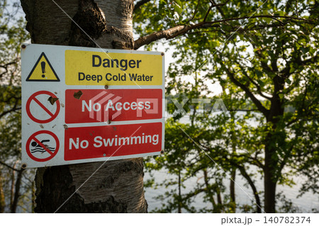 Danger sign warning of deep cold water. No access. No swimming. Warning sign on a tree with trees and lake behind. 140782374