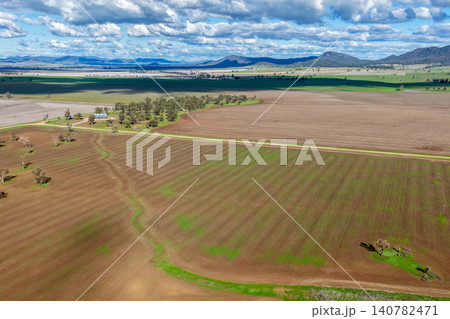 Aerial view of cultivated agricultural fields surrounded by lush green landscape, with a dirt road leading through the farmland, highlighting rural patterns and textures in NSW, Australia. Aerial view of cultivated agricultural fields surrounded by lush green landscape, with a dirt road leading through the farmland, highlighting rural patterns and textures in NSW, Australia. 140782471