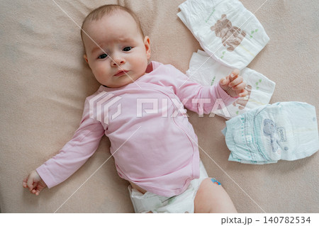 Close-up overhead shot of a sweet baby girl in pink clothes lying down on a soft blanket, surrounded by clean diapers during morning care. Adorable newborn infant lying on a neutral bed sheet with Close-up overhead shot of a sweet baby girl in pink clothes lying down on a soft blanket, surrounded by clean diapers during morning care. Adorable newborn infant lying on a neutral bed sheet with 140782534
