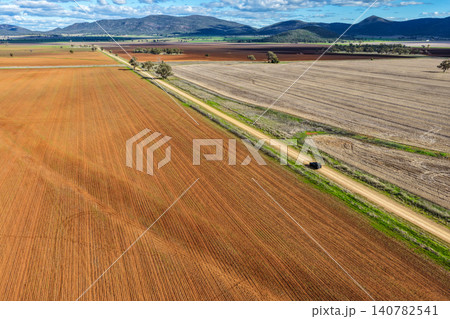 Aerial view of cultivated agricultural fields surrounded by lush green landscape, with a car travelling through dirt road leading through the farmland, highlighting rural patterns and textures. Aerial view of cultivated agricultural fields surrounded by lush green landscape, with a car travelling through dirt road leading through the farmland, highlighting rural patterns and textures. 140782541