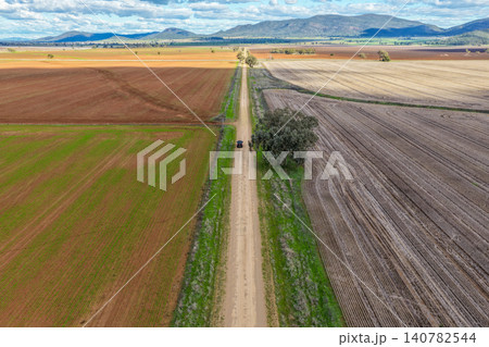Aerial view of a car travelling along a dirt road through cultivated agricultural fields in the Namoi Valley, with solitary trees and a mountain range creating a striking rural landscape. Aerial view of a car travelling along a dirt road through cultivated agricultural fields in the Namoi Valley, with solitary trees and a mountain range creating a striking rural landscape. 140782544