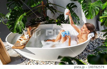 Happy young Caucasian woman in bubble bath pouring soap on sponge in modern green bathroom Happy young Caucasian woman in bubble bath pouring soap on sponge in modern green bathroom 140782574