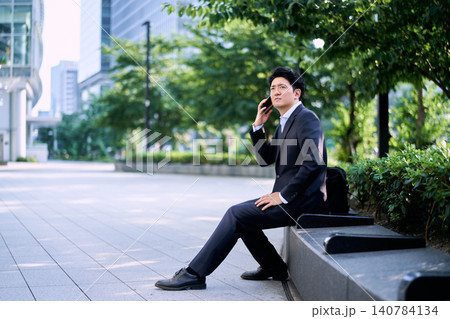 都会のオフィス街のベンチに座りスマートフォンで通話をする仕事中の30代の日本人の男性のビジネスマン 都会のオフィス街のベンチに座りスマートフォンで通話をする仕事中の30代の日本人の男性のビジネスマン 140784134