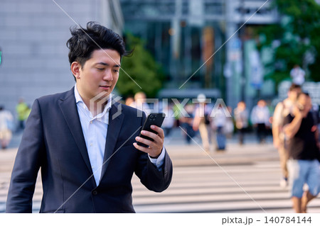 人々の行き交う夕方の都会のオフィス街の交差点の雑踏の中でスマートフォンを操作するビジネスマンの男性 140784144
