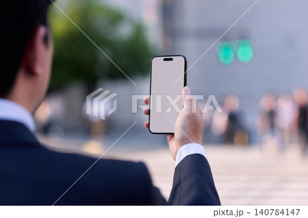 帰宅中に都会のオフィス街でスマートフォンを持ち画面を見て操作する顔無しの会社員の男性の後ろ姿と手元 140784147