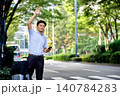 仕事の出張中にスマートフォンの配車のアプリで手配したタクシーを待つキャリーケースを持つ男性の会社員 140784283