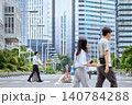 暑い夏の季節に都会のオフィス街の横断歩道をスーツケースを引いて歩くビジネスマンの男性と行き交う通行人 140784288