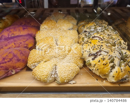 Selection of freshly baked gourmet pastries on a wooden display shelf. Featuring purple sweet potato buns, flower-shaped sesame bread and multigrain flower buns topped with pumpkin seeds. Selection of freshly baked gourmet pastries on a wooden display shelf. Featuring purple sweet potato buns, flower-shaped sesame bread and multigrain flower buns topped with pumpkin seeds. 140784486