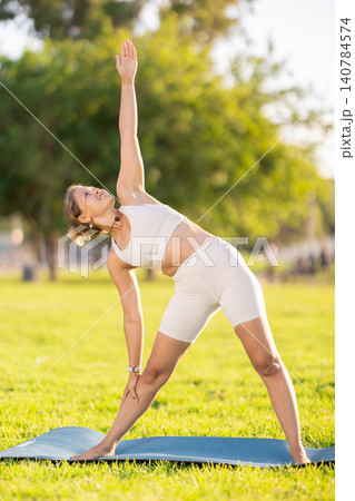 Girl in park on green grass of lawn performs Trikonasana, triangle asana 140784574