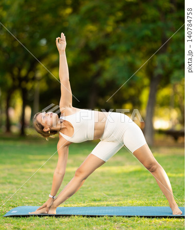 Girl in park on green grass of lawn performs Trikonasana, triangle asana 140784758
