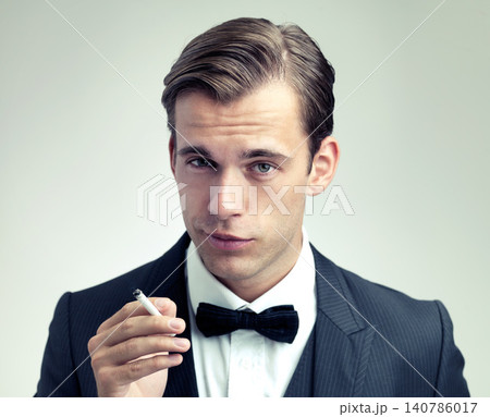 Portrait, elegant or young man smoking a cigarette in studio, formal or vintage fashion by white background. Face, attitude or gentleman in retro suit or tuxedo, bow tie or habit of tobacco at event Portrait, elegant or young man smoking a cigarette in studio, formal or vintage fashion by white background. Face, attitude or gentleman in retro suit or tuxedo, bow tie or habit of tobacco at event 140786017