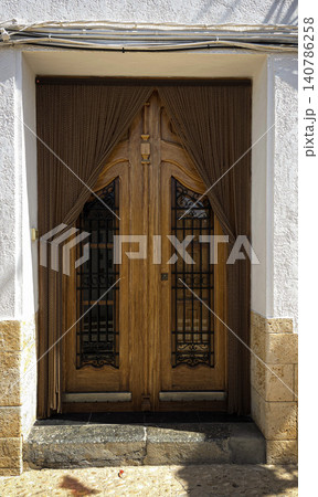 An ornate wooden double door features intricate iron grates. It has a decorative curtain hanging down. The door is set in a Mediterranean building with white walls. An ornate wooden double door features intricate iron grates. It has a decorative curtain hanging down. The door is set in a Mediterranean building with white walls. 140786258