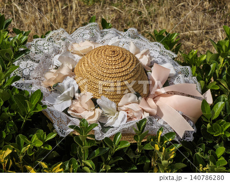 A vintage straw hat with lace, pink ribbons, and faux flowers lies on green bushes. The sun shines brightly in the garden, creating a warm atmosphere. A vintage straw hat with lace, pink ribbons, and faux flowers lies on green bushes. The sun shines brightly in the garden, creating a warm atmosphere. 140786280