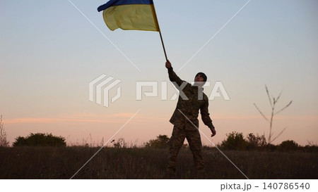 Male military waving blue-yellow banner on liberated territory occupied by russia. Young soldier of ukrainian army lifted flag of Ukraine in honor of victory against russian aggression at dusk Male military waving blue-yellow banner on liberated territory occupied by russia. Young soldier of ukrainian army lifted flag of Ukraine in honor of victory against russian aggression at dusk 140786540
