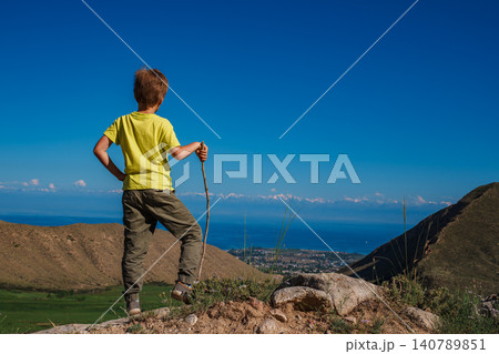 Boy hiker with stick looks at the lake shore from the top of the mountain Boy hiker with stick looks at the lake shore from the top of the mountain 140789851
