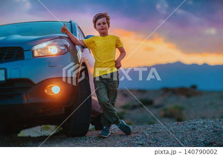 Successful boy standing by the car and showing thumbs up at twilight on mountains background 140790002